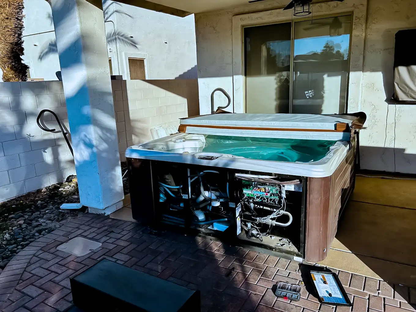 Spa equipment compartment open with controls and wiring exposed beside a patio